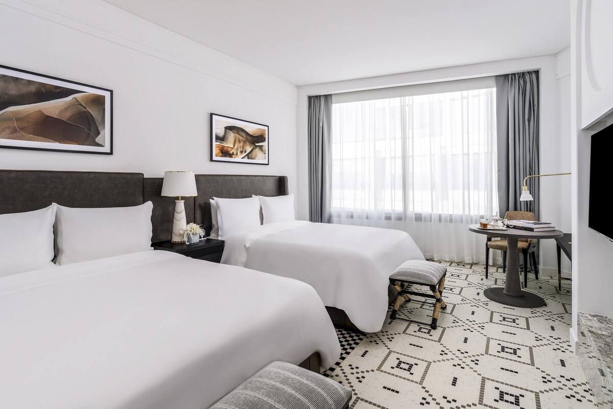 A hotel room with two beds, a desk with a chair, a TV, and artwork on the walls.