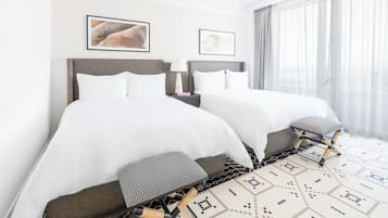A hotel room with two beds, a nightstand, a lamp, and a view of the city through large windows.