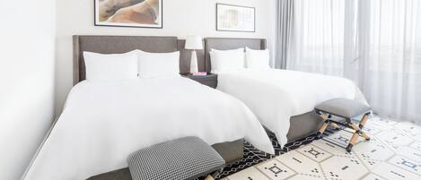 A hotel room with two beds, a nightstand, a lamp, and a view of the city through large windows.