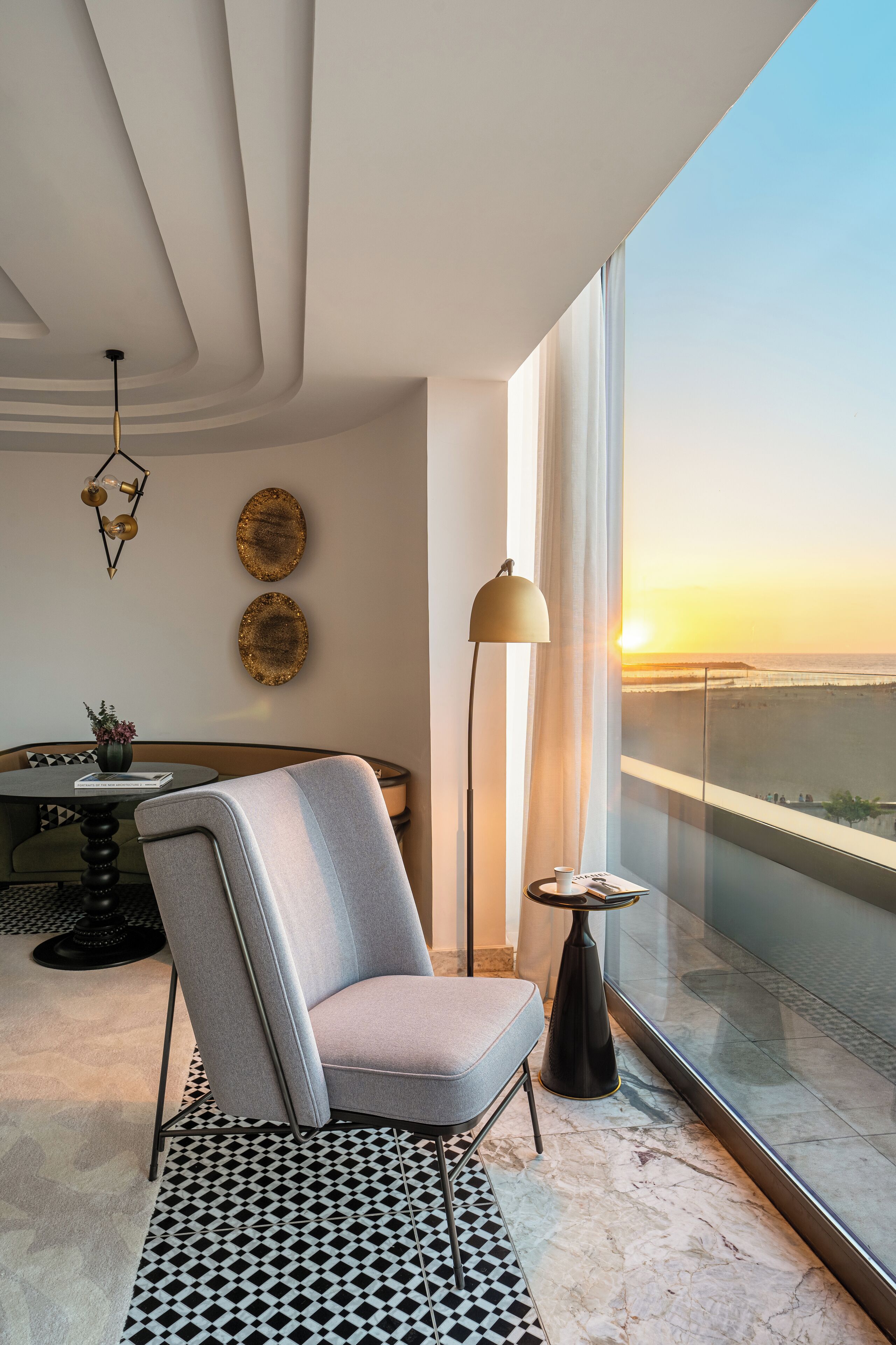 Ein modernes Wohnzimmer mit Blick auf den Sonnenuntergang, ein runder Tisch mit einer Vase, ein grauer Sessel und eine Stehlampe.
