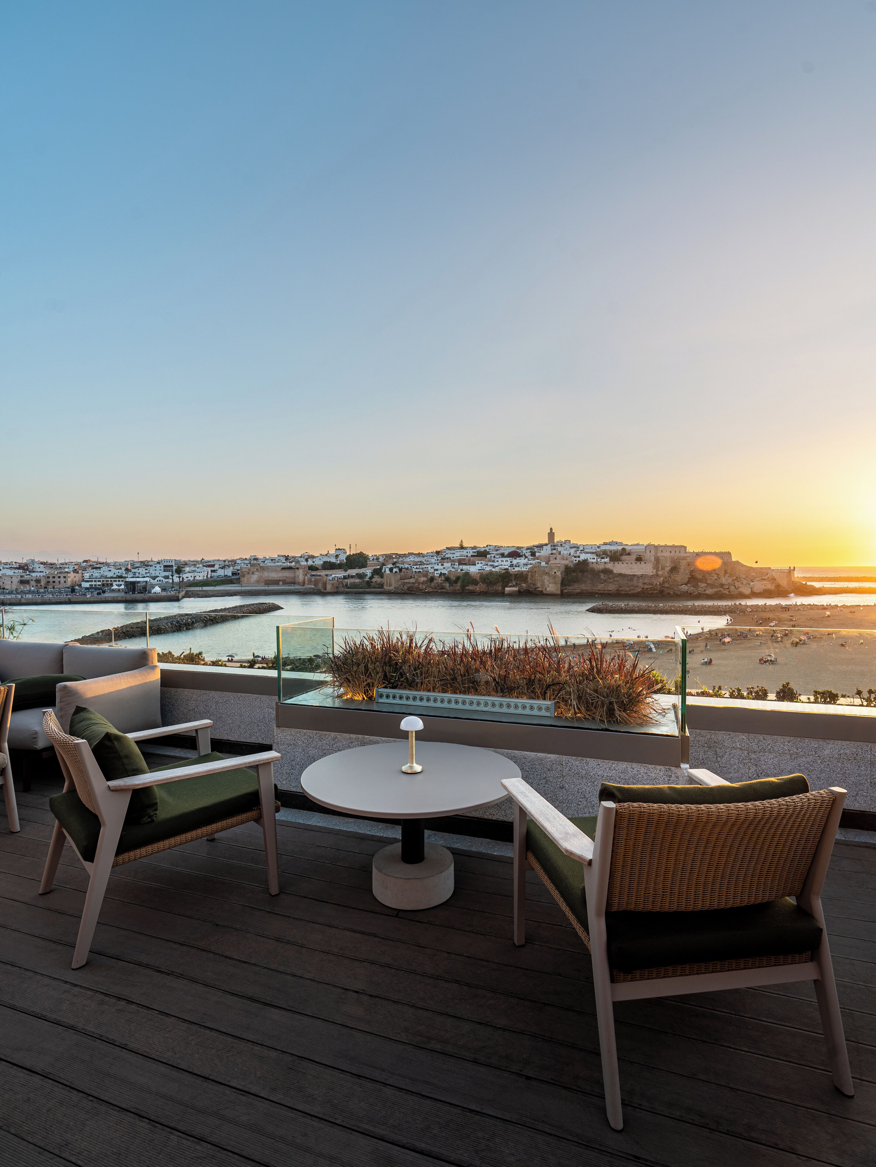 A terrace with wicker chairs and a table, overlooking a coastal cityscape during sunset.