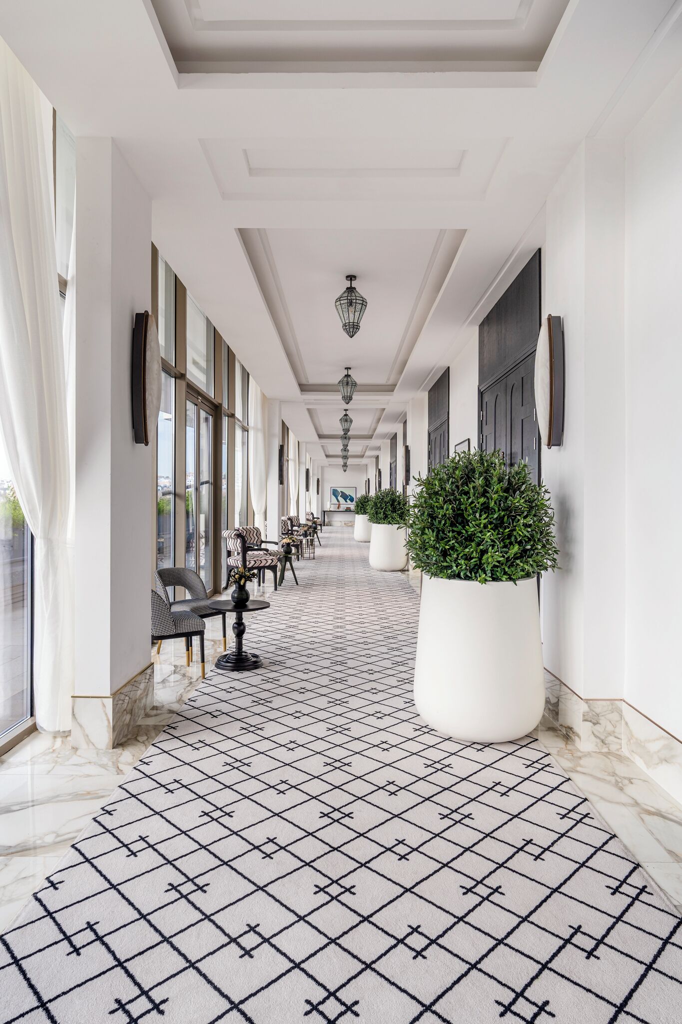 A hotel corridor with a patterned floor, seating areas, large windows, and potted plants.