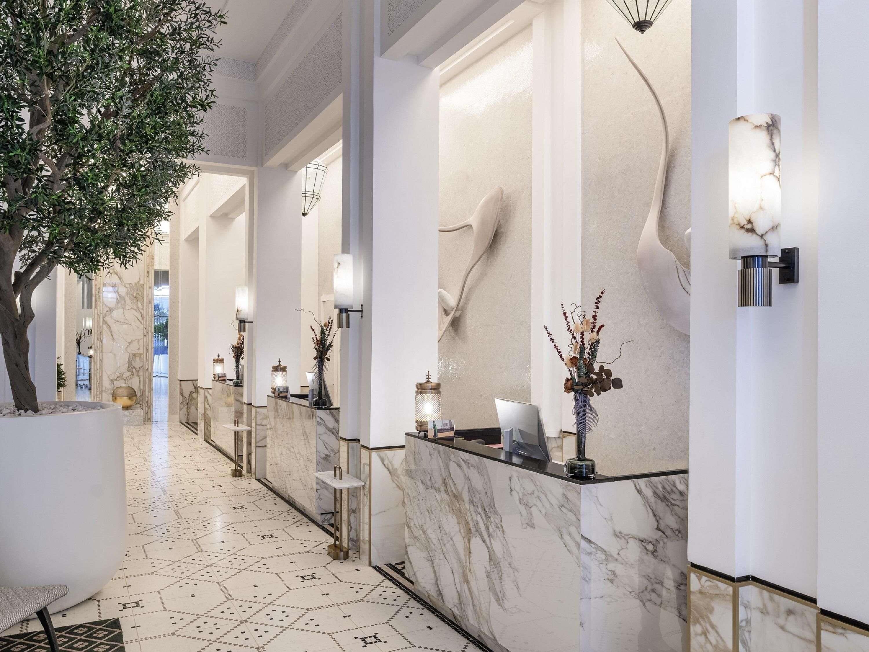 A hotel lobby with a marble floor, a reception desk, a large tree, and wall-mounted lighting.