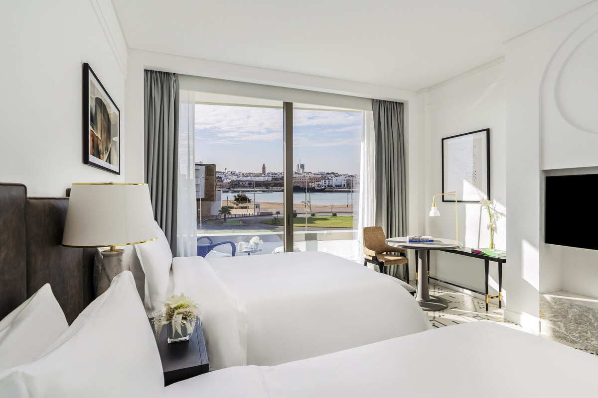 A hotel room with a large bed, a desk, a chair, a TV, and a view of the city and water.