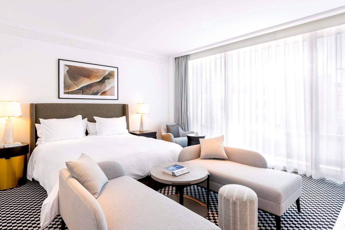 A hotel room with a bed, two armchairs, a small table, and a large window with sheer curtains.