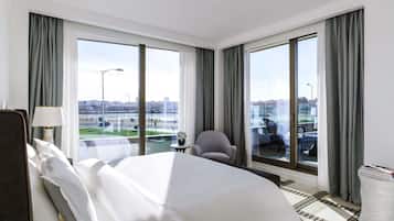 A hotel room with a large bed, a chair, a table, and a view of the city through large windows.