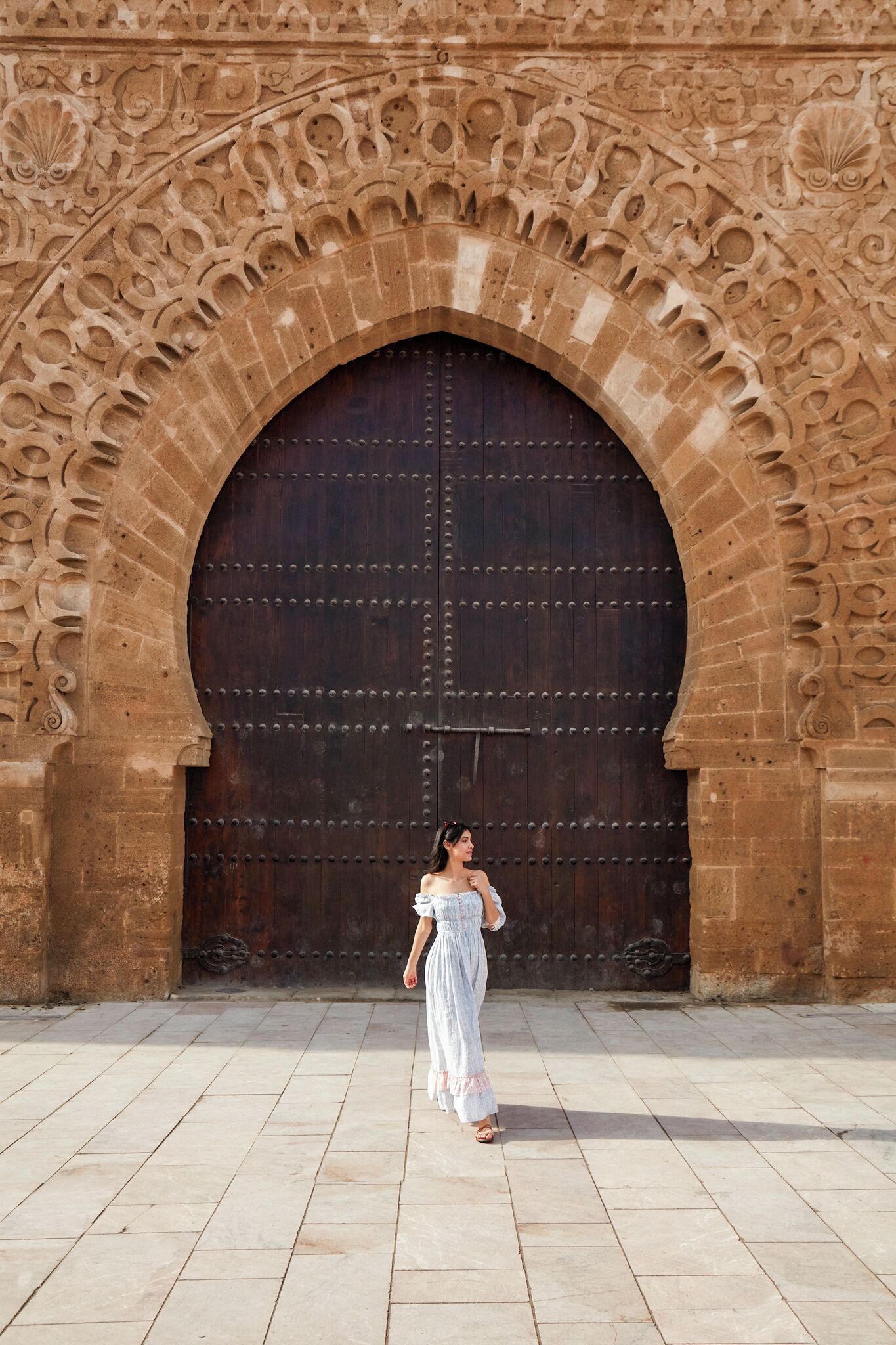 A woman walking in front of a large, ornate stone archway with a heavy wooden door.
