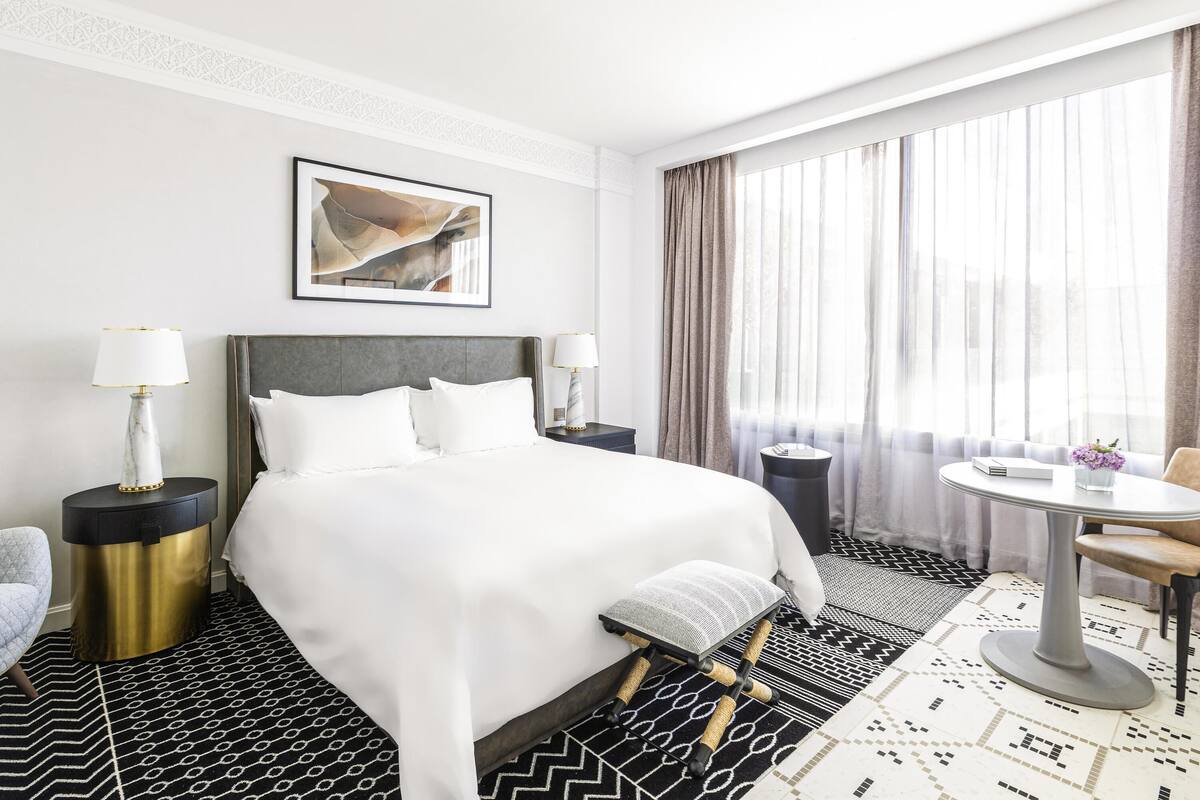 A hotel room with a bed, two bedside tables, a chair, a round table, and a window with curtains.