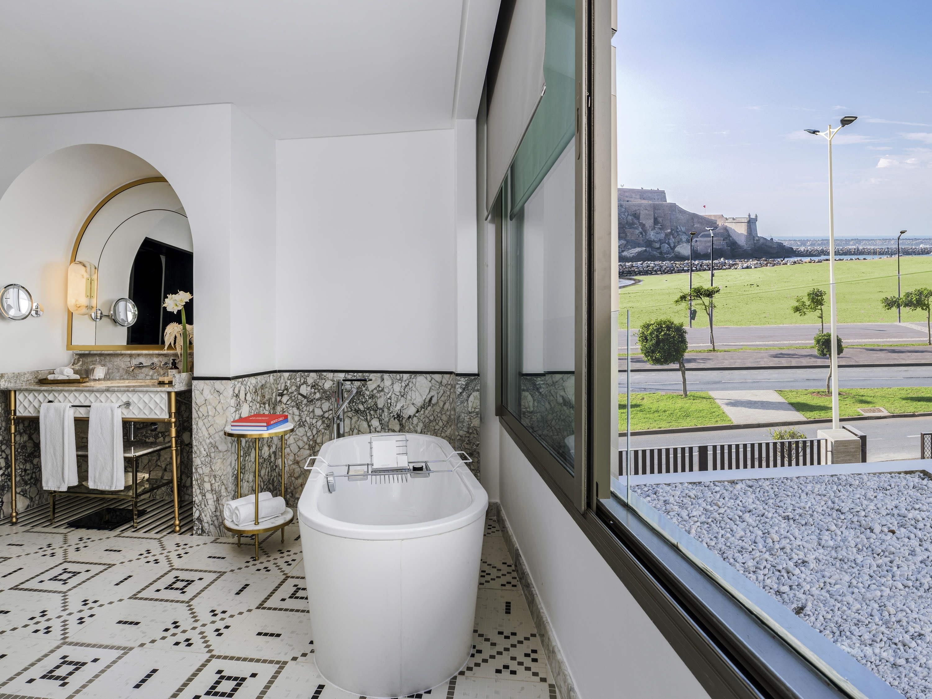 Une salle de bain avec une baignoire, un meuble-lavabo avec un miroir et une vue sur un château par la fenêtre.