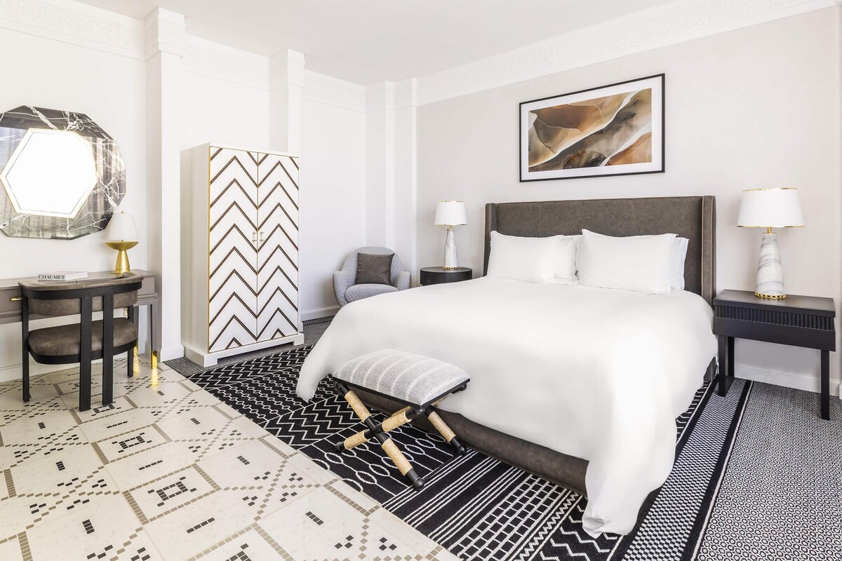 A hotel room with a bed, a desk, a chair, a nightstand, a lamp, a mirror, and a wall art piece.