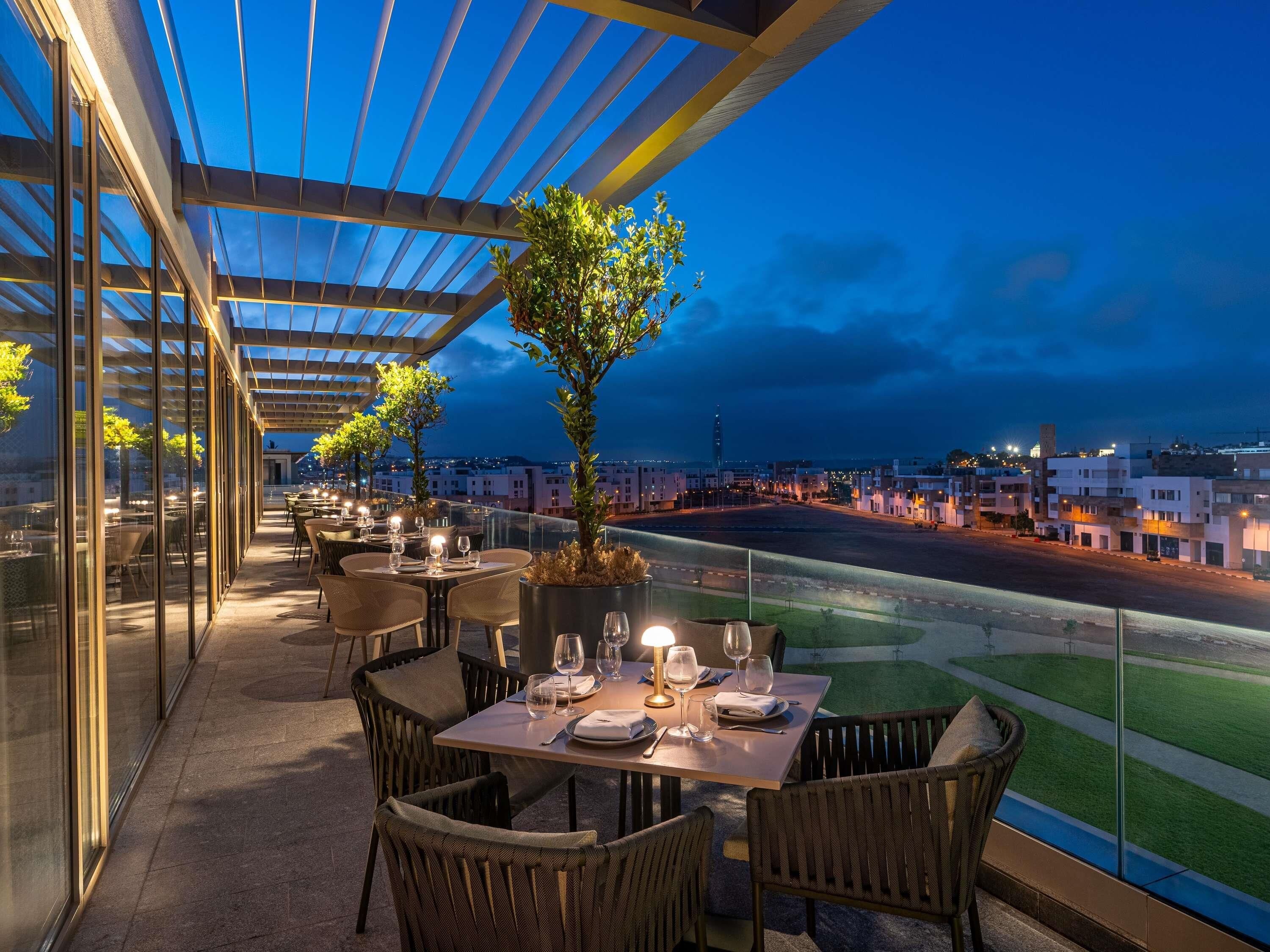 Un espace extérieur aménagé pour les repas, avec des tables et des chaises, une rambarde en verre et une vue sur le paysage urbain.