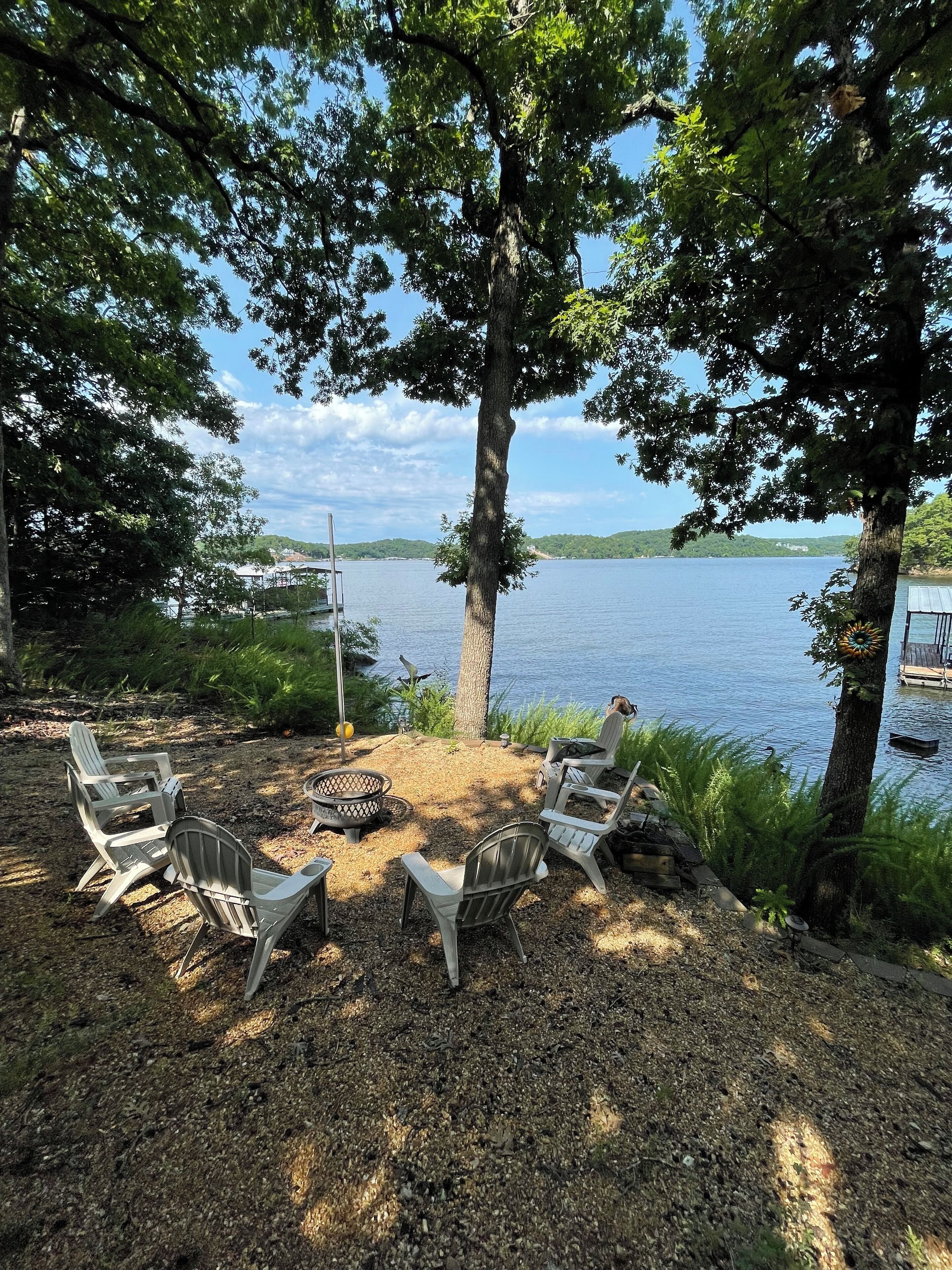 A lakeside seating area with Adirondack chairs and a fire pit, surrounded by trees and a view of the lake.