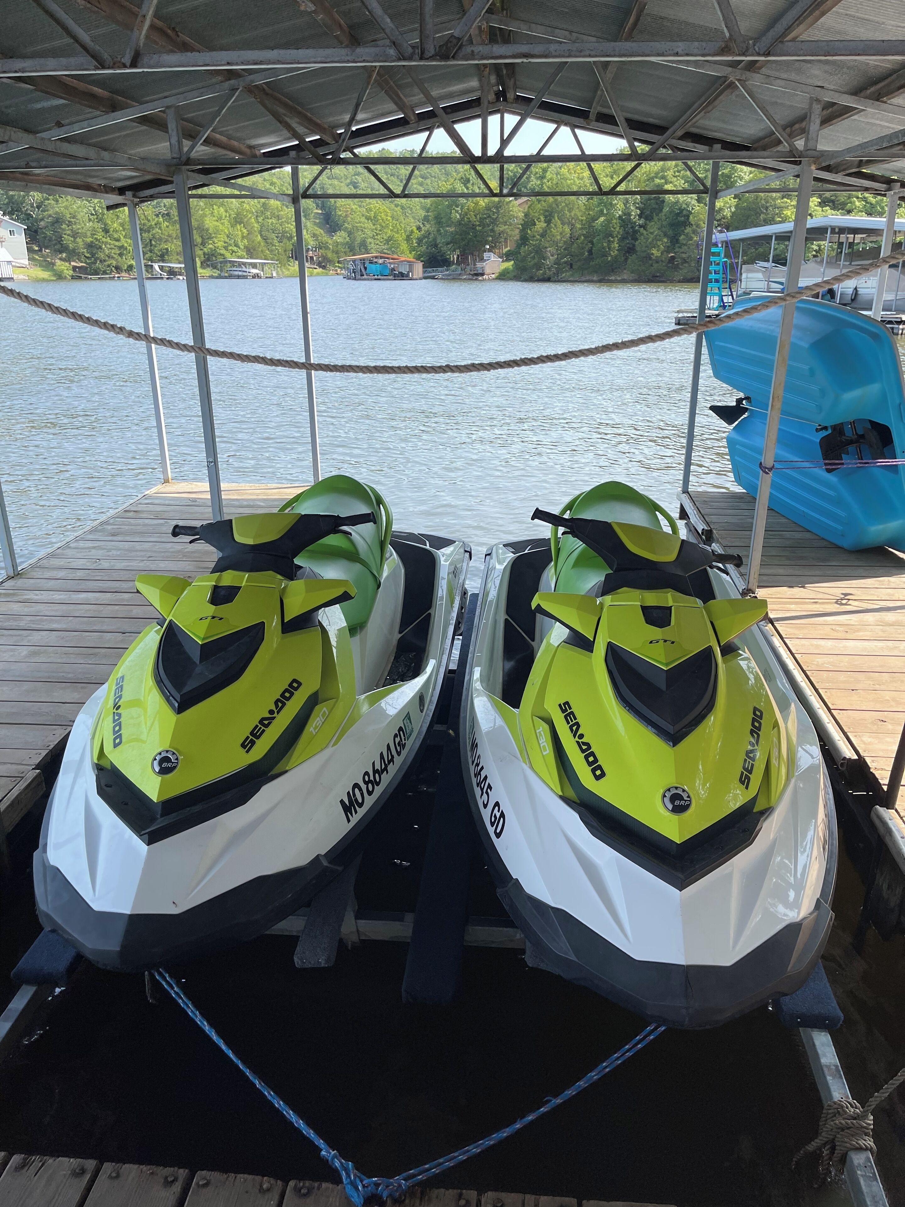 Deux jet skis Sea-Doo sont garés sur un quai en bois, à l’abri sous une structure couverte, d’où l’on peut admirer un lac et des bateaux au loin.