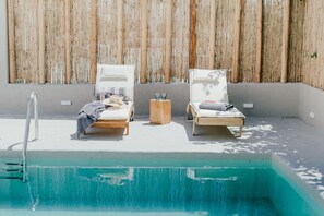 A poolside area with two lounge chairs, a small wooden stool, and a metal ladder leading into the pool.