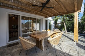 An outdoor patio with a wooden table and wicker chairs, a ceiling fan, and a stone floor.