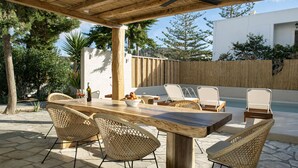 An outdoor patio with a wooden table and wicker chairs, a pool, and a white building in the background. - Condo with private pool - 5 minutes from the beach (Pollonia)