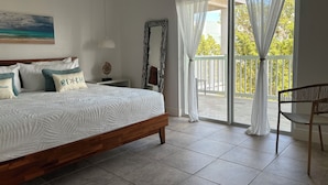 Ein Schlafzimmer mit einem großen Bett, einem Nachttisch, einem Gemälde, einem Sessel und Blick ins Freie durch eine Glastür.