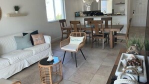 Ein modernes Wohnzimmer mit einem weißen Sofa, einem Holzespeisesaal und Stühlen. Im Hintergrund befindet sich eine Küche mit weißen Schränken und einer Chandelier.