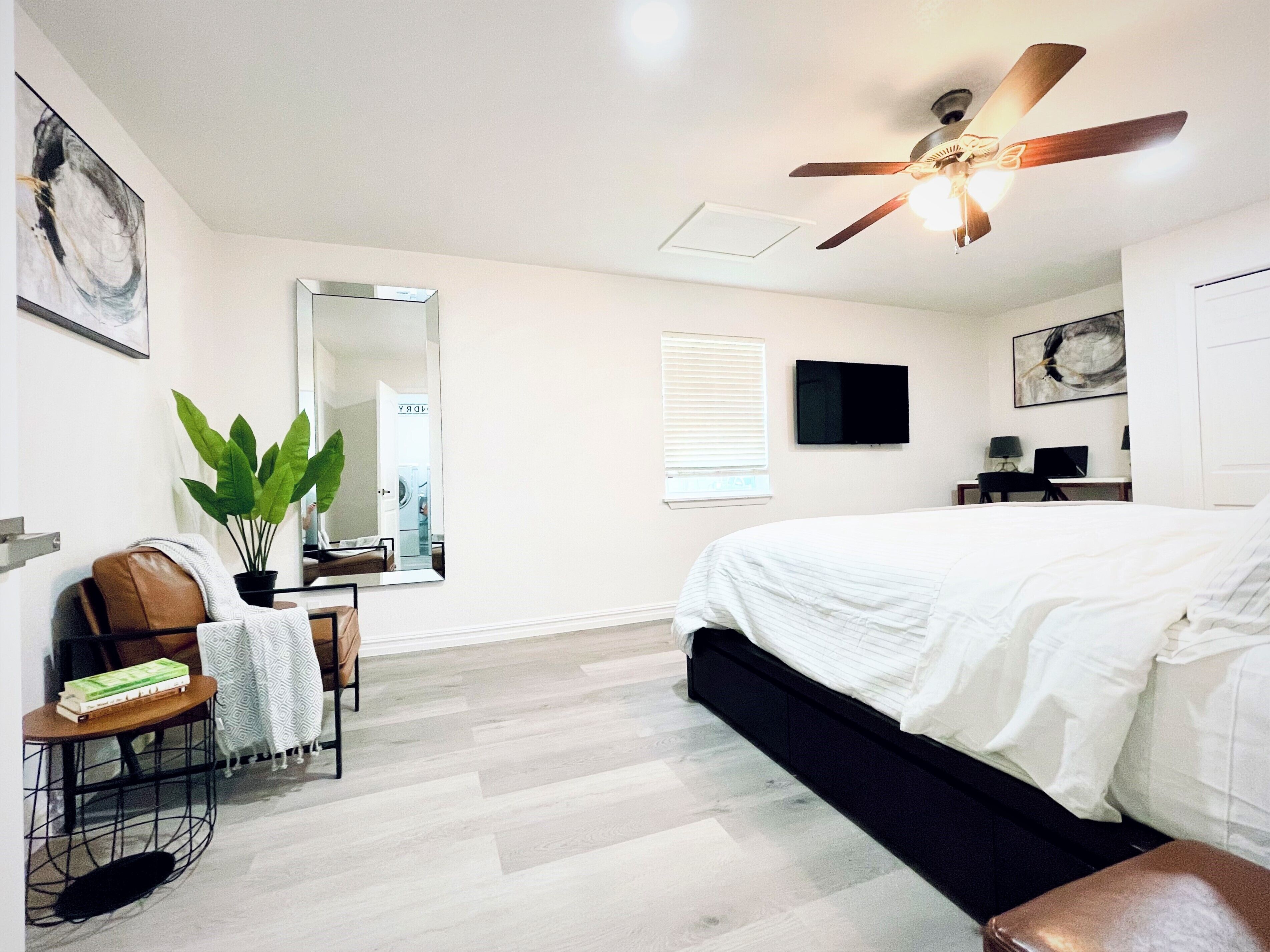 A bedroom with a bed, a ceiling fan, a large mirror, a potted plant, a desk with a chair, and a television on the wall.