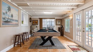 Una mesa de billar en una habitación con una base de madera en forma de X, taburetes de madera y vistas al exterior a través de amplios ventanales.