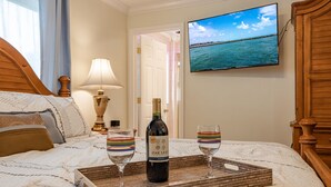 Una cama bien hecha con una botella de Oak Leaf Merlot y dos copas sobre una bandeja, un televisor mostrando una escena costera y un cabecero de madera.