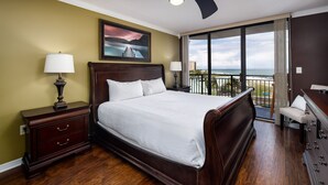 A hotel room with a large bed, a nightstand, a dresser, a balcony with a view of the ocean, and a painting on the wall.