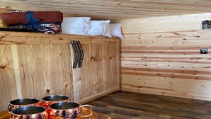 Interior de una cabaña de madera con una cama, almohadas, un armario de madera y tazas de cobre sobre una bandeja.