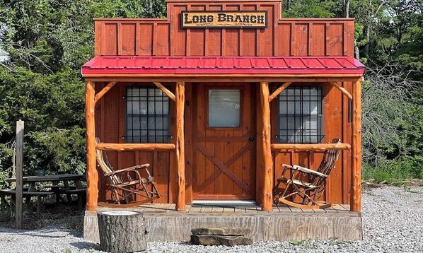 A rustic wooden building with a red roof, a sign reading "Long Branch," and two rocking chairs outside.