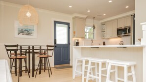 A modern kitchen with white cabinets, a blue door, and a dining area with wooden chairs and white stools. - Wingspan: Modern, Elegant Home Close to the Intracoastal Waterway (Oak Island)