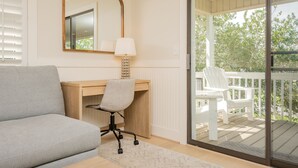 A modern bedroom with a grey sofa, a wooden desk with a chair, a lamp, and a large mirror. There is a sliding glass door leading to an outdoor deck with white chairs. - Wingspan: Modern, Elegant Home Close to the Intracoastal Waterway (Oak Island)