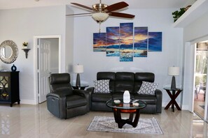 Una sala de estar con un ventilador de techo, un sofá de cuero negro, dos sillones, una mesa de centro y un cuadro grande de un paisaje en la pared.