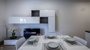 Un moderno comedor con una mesa blanca dispuesta para seis personas, incluyendo platos, vasos y cubiertos. Hay una silla blanca a la izquierda y un pequeño armario blanco sobre la mesa.