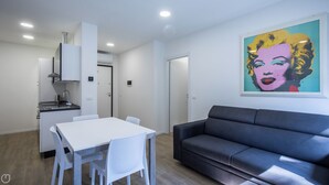 A modern living room with a black sofa and a dining table with chairs. There is a kitchen area with a stove and cabinets. A colorful artwork is on the wall. - Vat 52 Sub 15 - One Bedroom Apartment, Maximum 4 Guests (Udine)