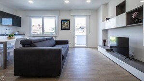 A modern living room with a grey sofa, a flat-screen TV on a wooden stand, and a kitchen area with a sink and cabinets. - Vat 52 Sub 16 - One Bedroom Apartment, Maximum 5 Guests (Udine)
