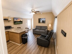 A compact living space with a kitchenette, a gray sofa, a black leather chair, and a ceiling fan. - Rural Getaway. Sleeps 2, Near Paul B State Park (Hattiesburg)