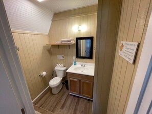 Baño pequeño con inodoro, lavamanos y espejo. Hay un estante sobre el lavamanos y un cartel en la pared.