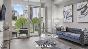 A modern living room with a glass coffee table, a grey sofa, and a city view through large sliding doors.