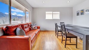 A modern living room with a leather sofa, wooden dining table, and chairs. A ceiling fan is visible, and there are large windows offering a mountain view.