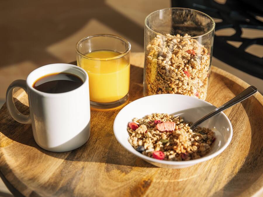 A wooden table with a white mug, a glass of orange juice, a glass of coffee, and a bowl of granola.