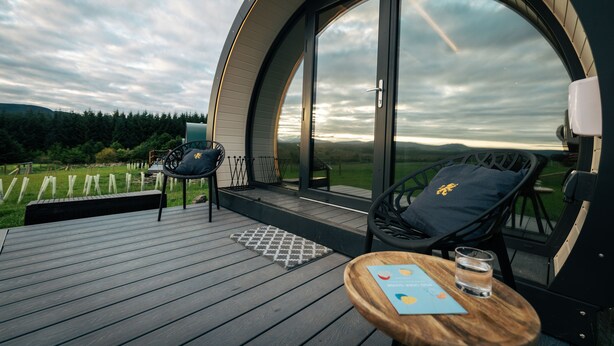 A circular room with a large window, a wooden table with a glass, and two chairs with cushions. - Cardross Estate Glamping (Stirling)