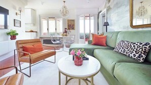Uma sala de estar espaçosa com um sofá verde, uma poltrona de couro e uma mesa de centro. Há duas cadeiras, um candelabro e um espelho grande na parede.