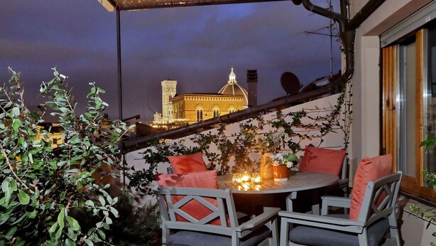 Um pátio externo com duas cadeiras cinzas e uma mesa, rodeado por vegetação e vista para um edifício iluminado ao anoitecer.