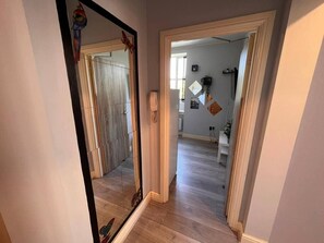 A hallway with a wooden floor, a mirror, and a door leading to a room with a white wall and a window.