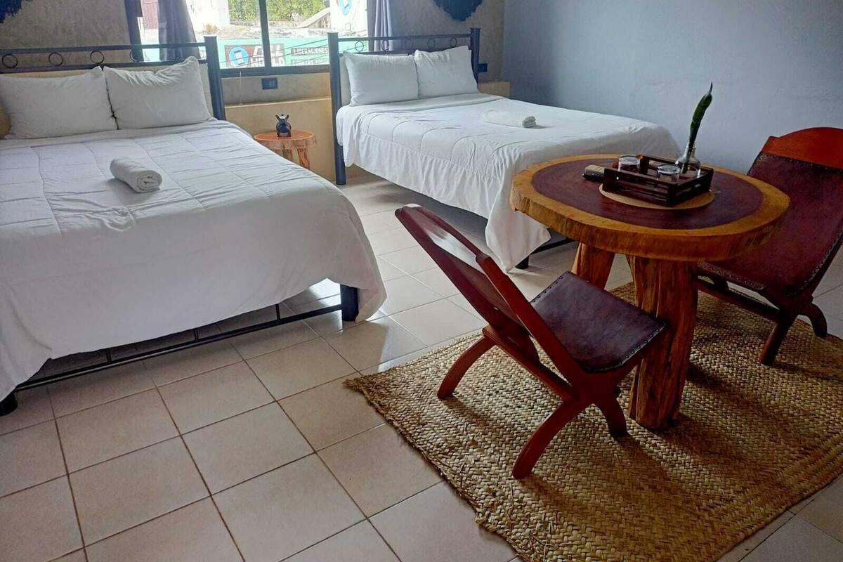 Habitación de hotel con dos camas, una mesita redonda con una silla y vistas al exterior a través de una ventana.
