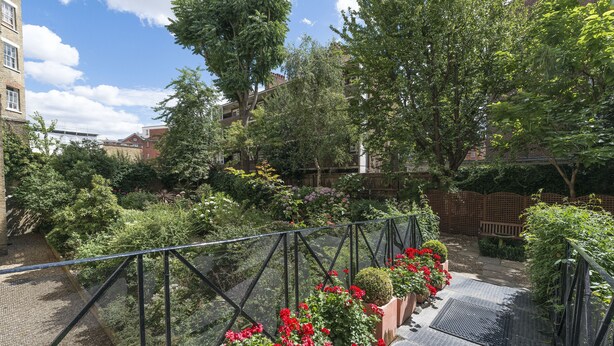 A rooftop garden with a metal railing, potted plants, and a paved walkway.