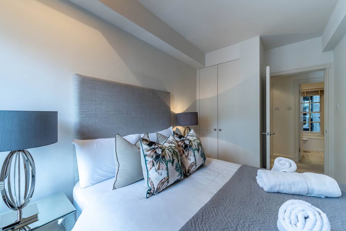 A modern hotel room with a bed, pillows, a bedside table with a lamp, and a door leading to another room.