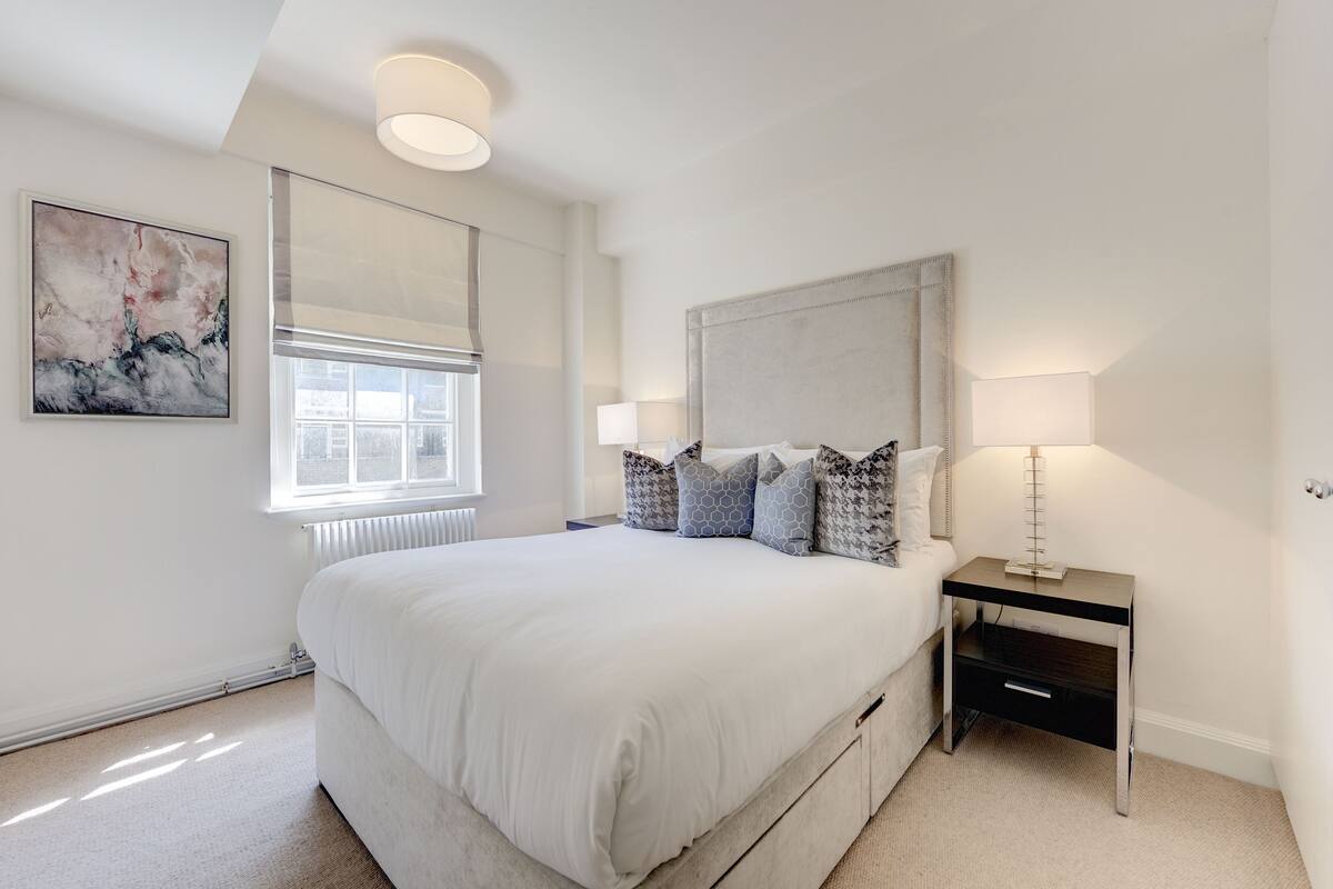A bedroom with a large bed, a nightstand with a lamp, a window with blinds, and a wall-mounted artwork.