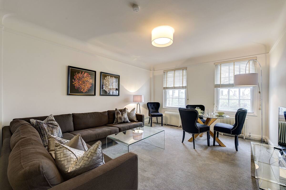 A living room with a brown sofa, a glass coffee table, a dining table with chairs, and artwork on the walls.