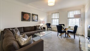 A living room with a brown sofa, a glass coffee table, a dining table with chairs, and artwork on the walls.