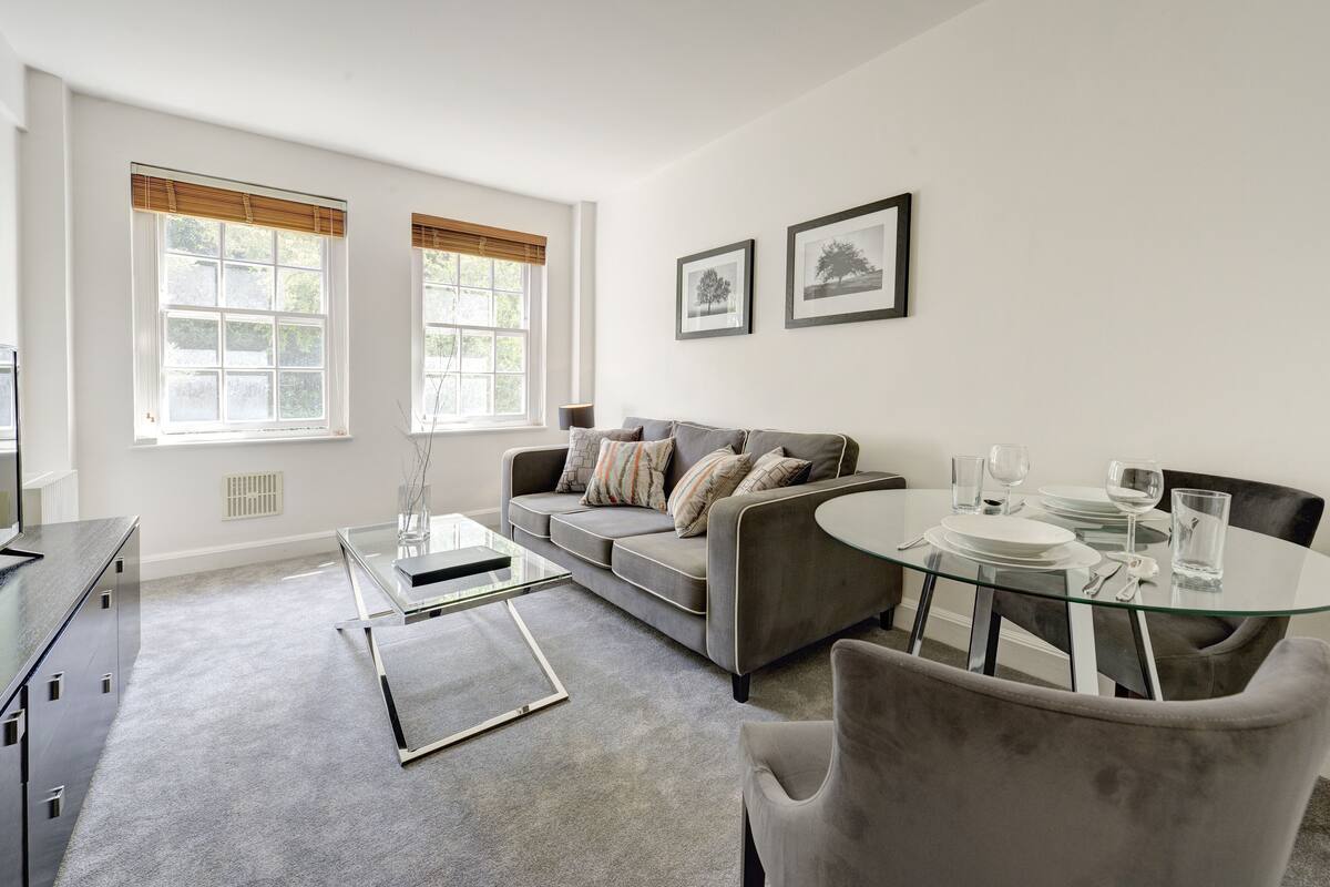 A modern living room with a grey sofa, a glass coffee table, and a dining table set for two.
